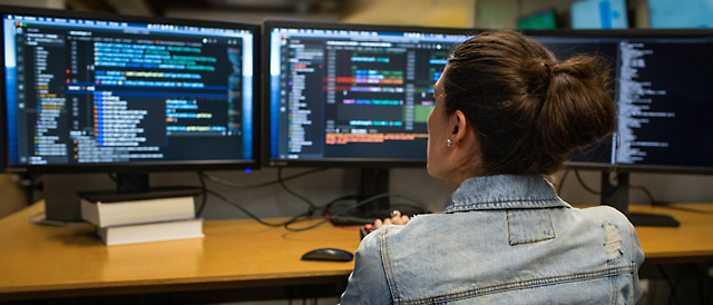 在光线昏暗的办公室里，一名女子在显示软件代码的多个计算机屏幕上编写代码。