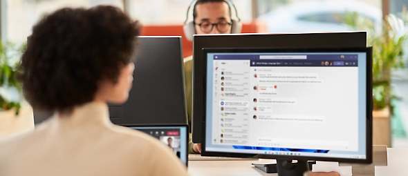 Donna che collabora durante una riunione di Microsoft Teams mentre lavora in un ambiente di ufficio aperto su un monitor doppio